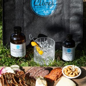 Clive's reusable picnic bag with a logo, containing two bottled cocktails and a charcuterie board, displayed on the grass